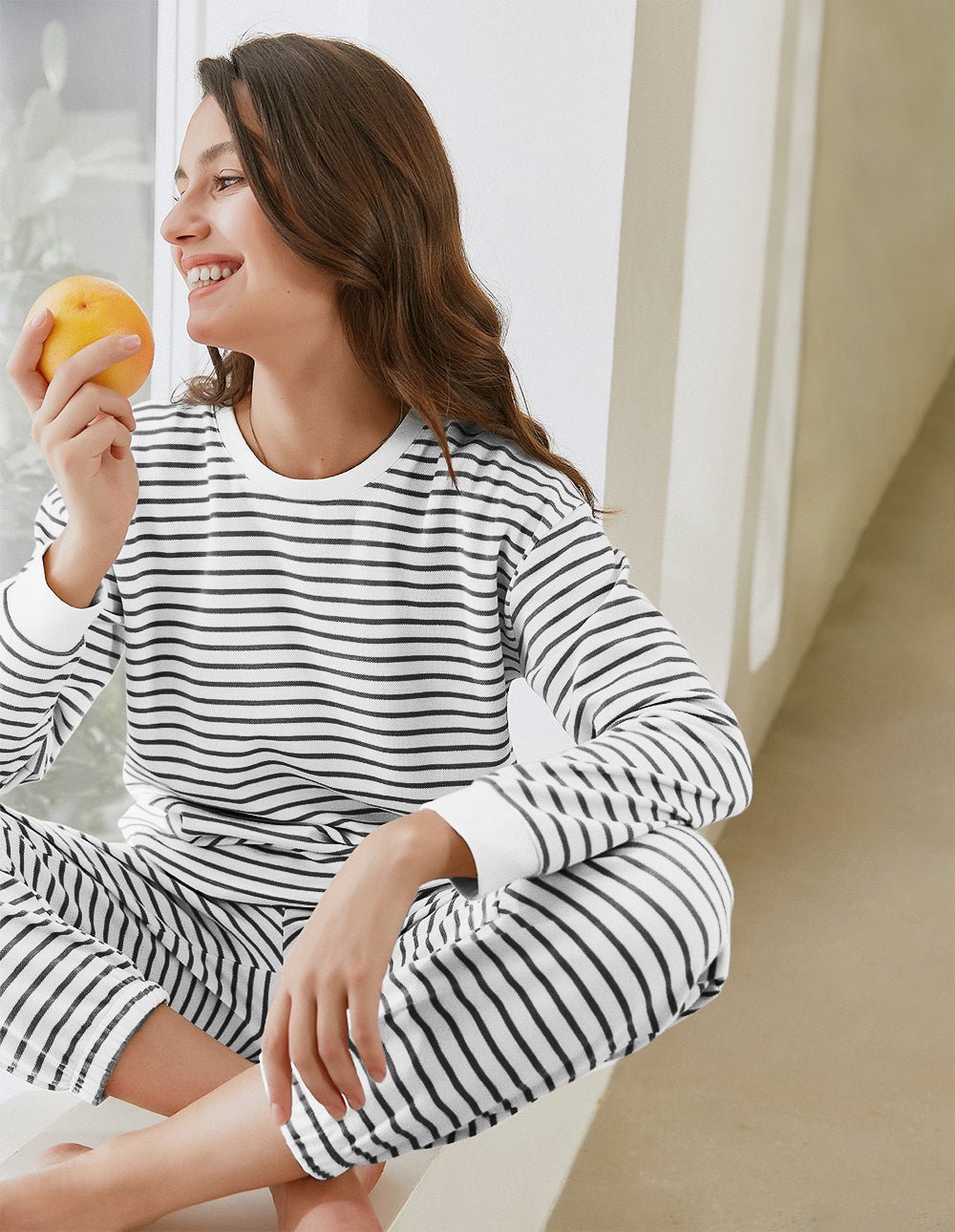 Striped cotton pajama set, long sleeves and pants, cream with tan stripes, seated and holding orange